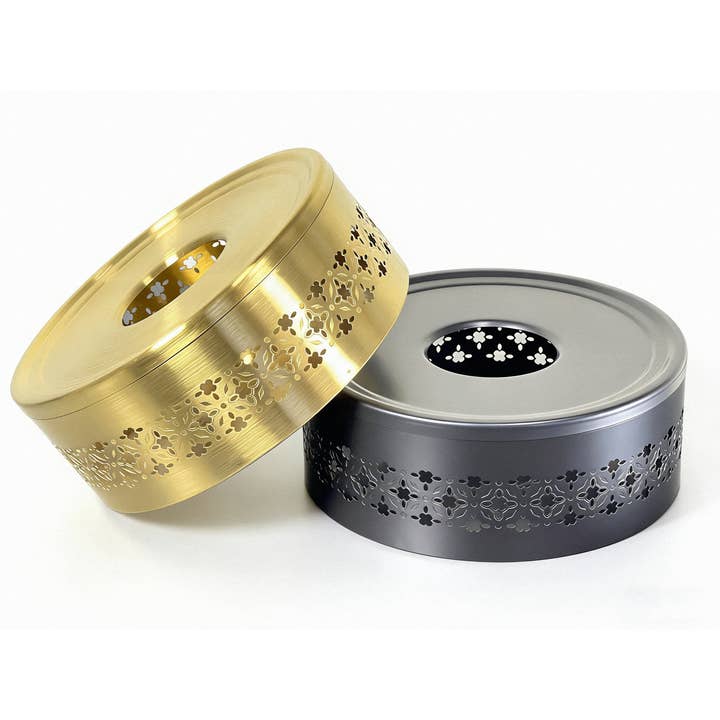 Hana Tea Warmer in Black with Hana tea warmer in gold resting on top, both with decorative detailing, designed to maintain tea temperature using a standard tealight candle.