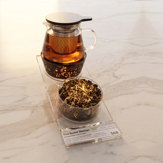 Tea Scent Station tea set on a clear acrylic stand, featuring a glass teapot with infuser and metal tea warmer, a Aroma Scent Cup filled with loose-leaf tea, and a small acrylic label plate with printed price/brand card.