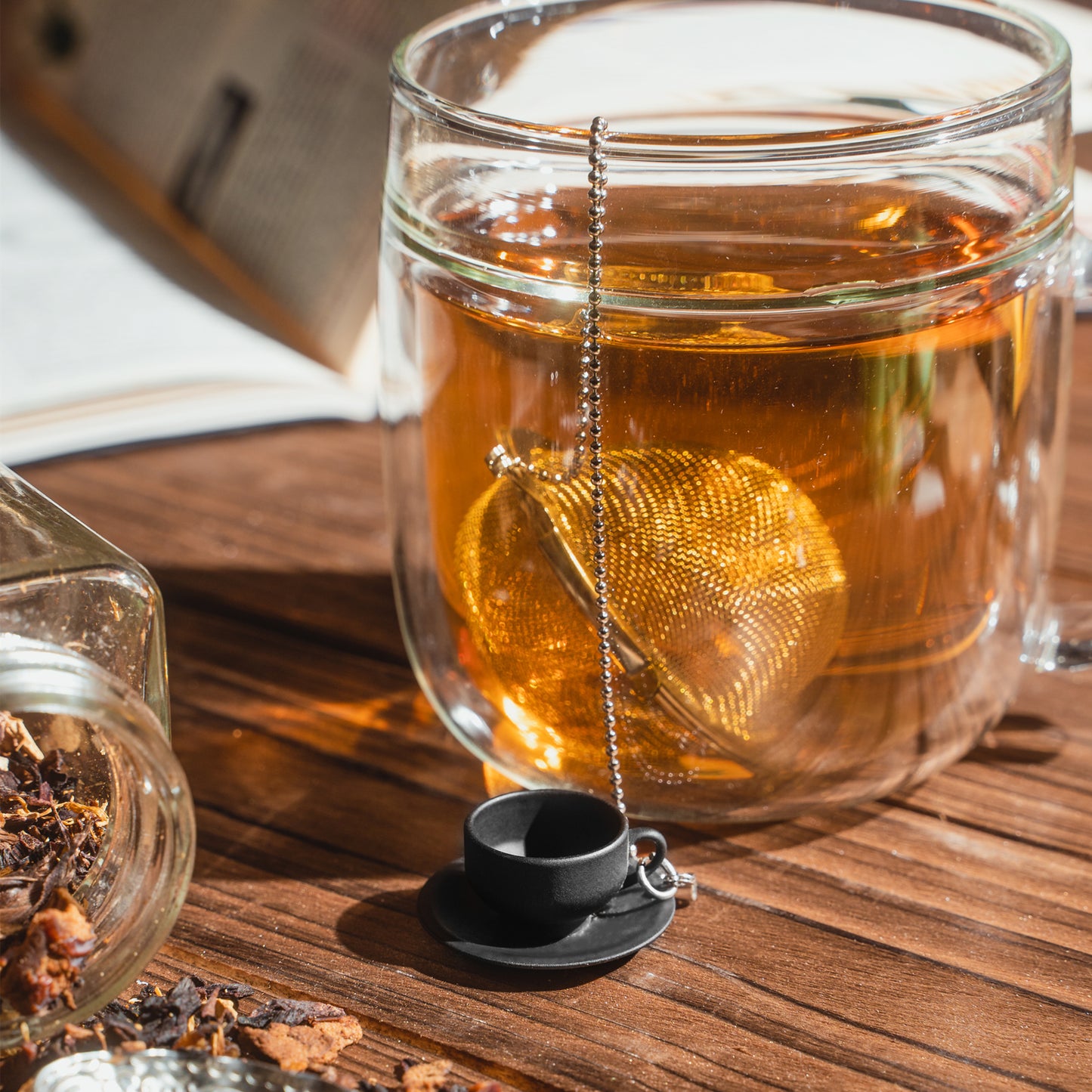 01Living ChiChi Loose Leaf Tea Ball Infuser sitting next to a cup of steeped tea, with its decorative cute tea cup charm and chain adding a playful touch to the tea ritual.