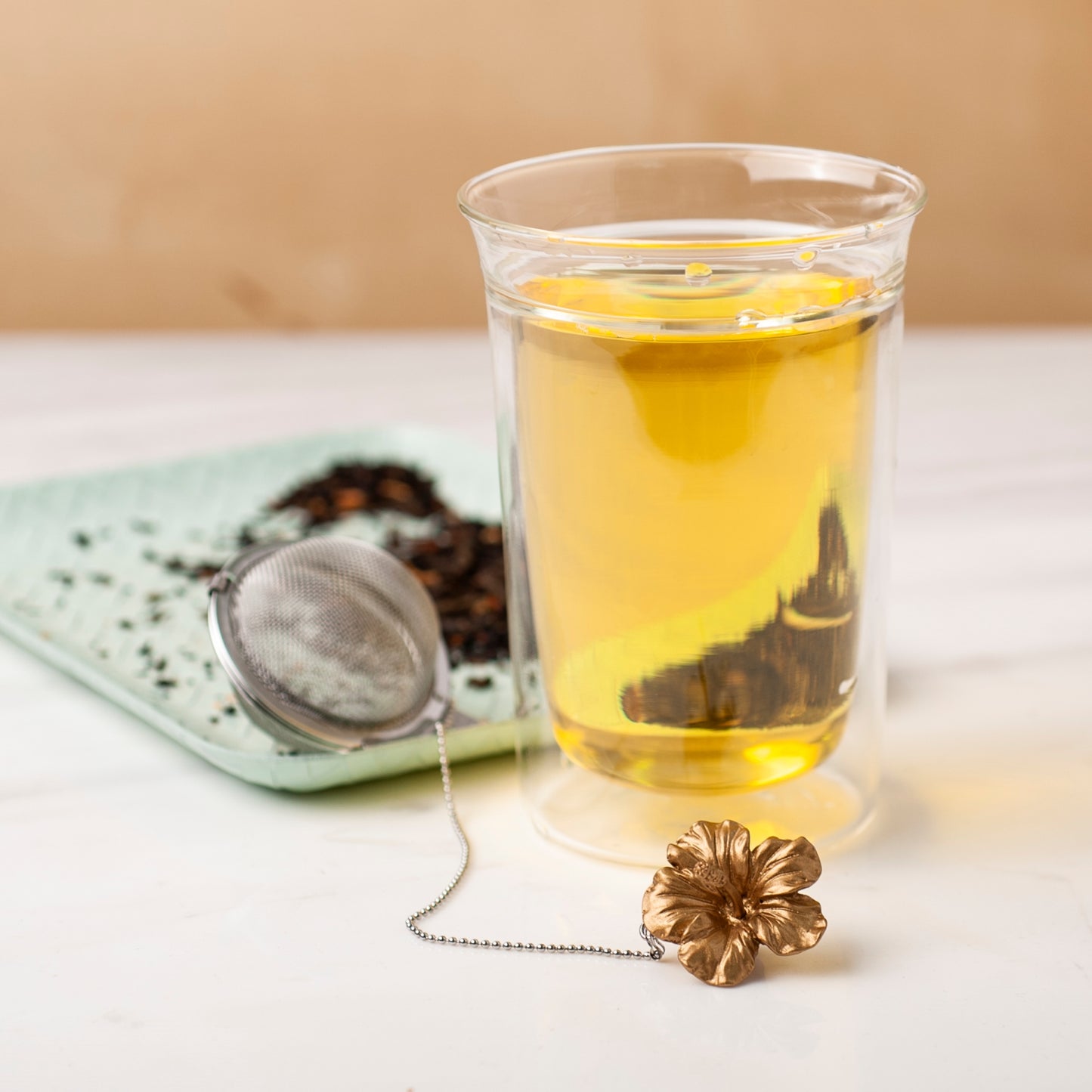 01Living ChiChi Loose Leaf Tea Ball Infuser sitting next to steeped tea in a cup, with its decorative charm and chain adding a playful touch to the tea ritual.