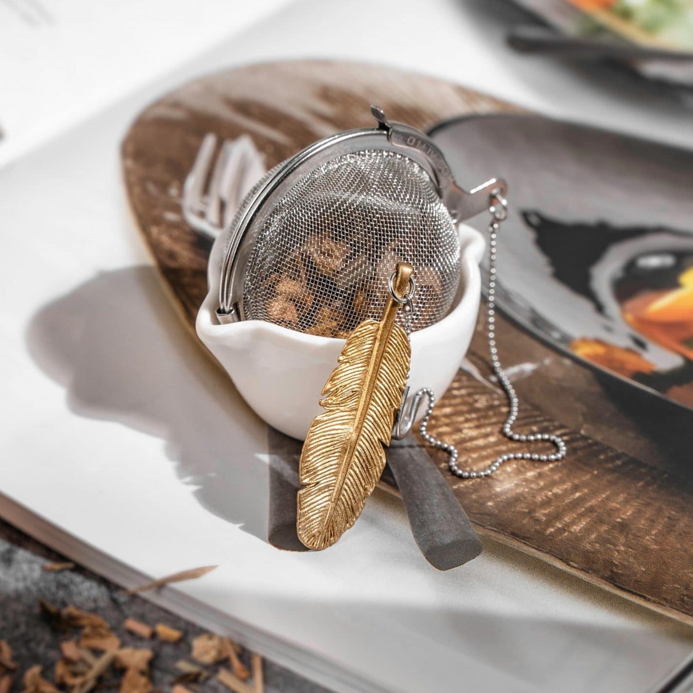 Loose Leaf Tea Ball Infuser crafted from 304 food-grade stainless steel, filled with loose leaf tea in a fine mesh, attached chain and decorative Gold Feather charm for easy loose-leaf tea brewing.