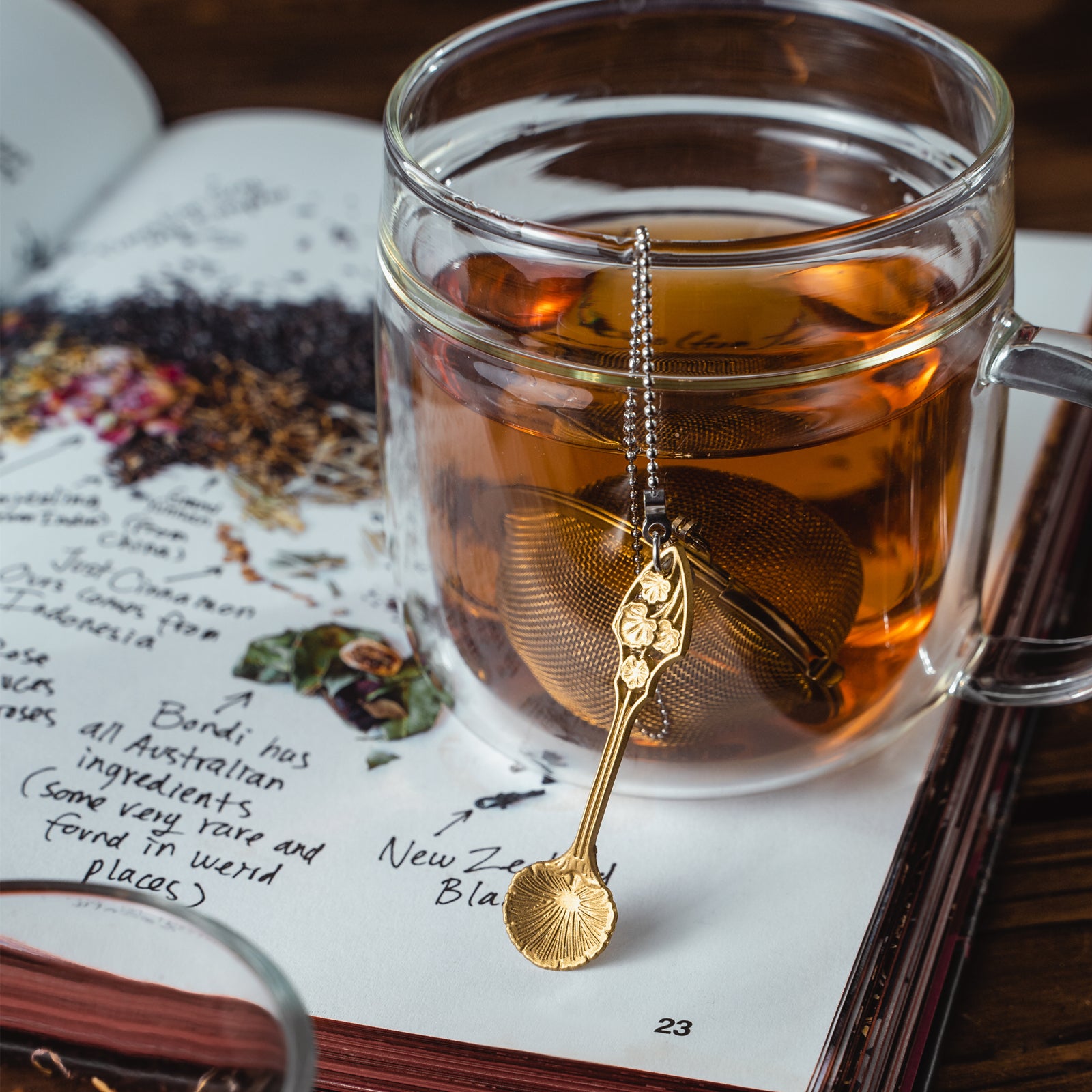 Stainless steel Loose Leaf Tea Ball Infuser with a gold Lobed Spoon Charm, filled with loose-leaf tea and suspended in a cup, allowing tea leaves to steep while keeping sediment contained.
