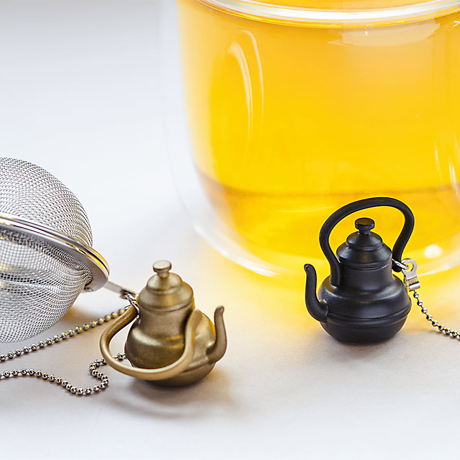 Stainless steel Loose Leaf Tea Ball Infusers with gold and black tall teapot charms, sitting alongside a brewed cup of tea.