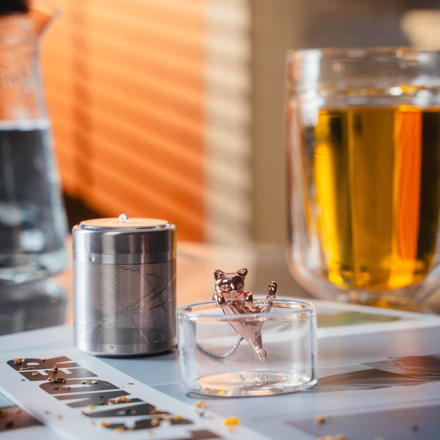 01Living stainless steel tea infuser with a rose gold cat-shaped ChiChi ornament, set on a dark wooden surface with dried tea ingredients and a glass of brewed tea in background