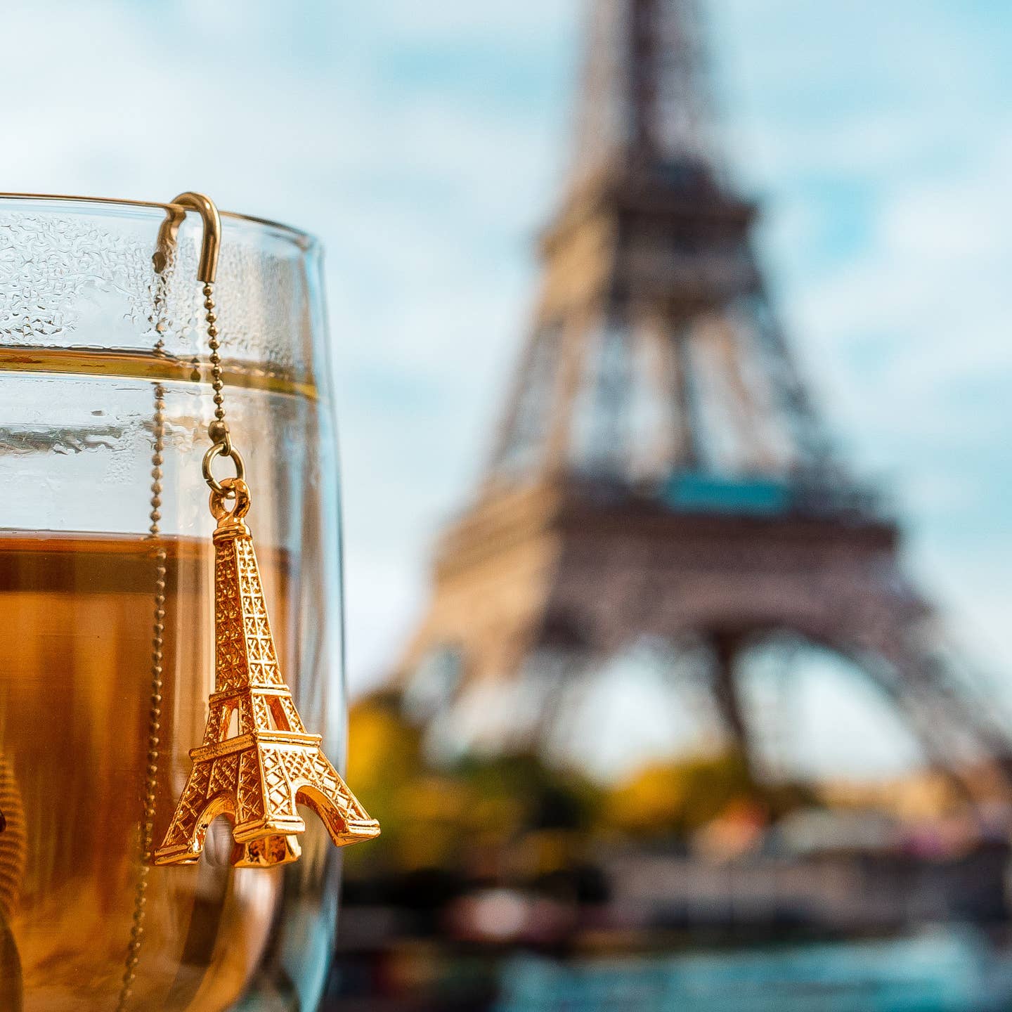 01Living tea infuser with gold Eiffel Tower pendant ChiCHi charm hanging from a clear glass of brewed tea, with a blurred Eiffel Tower in the background.