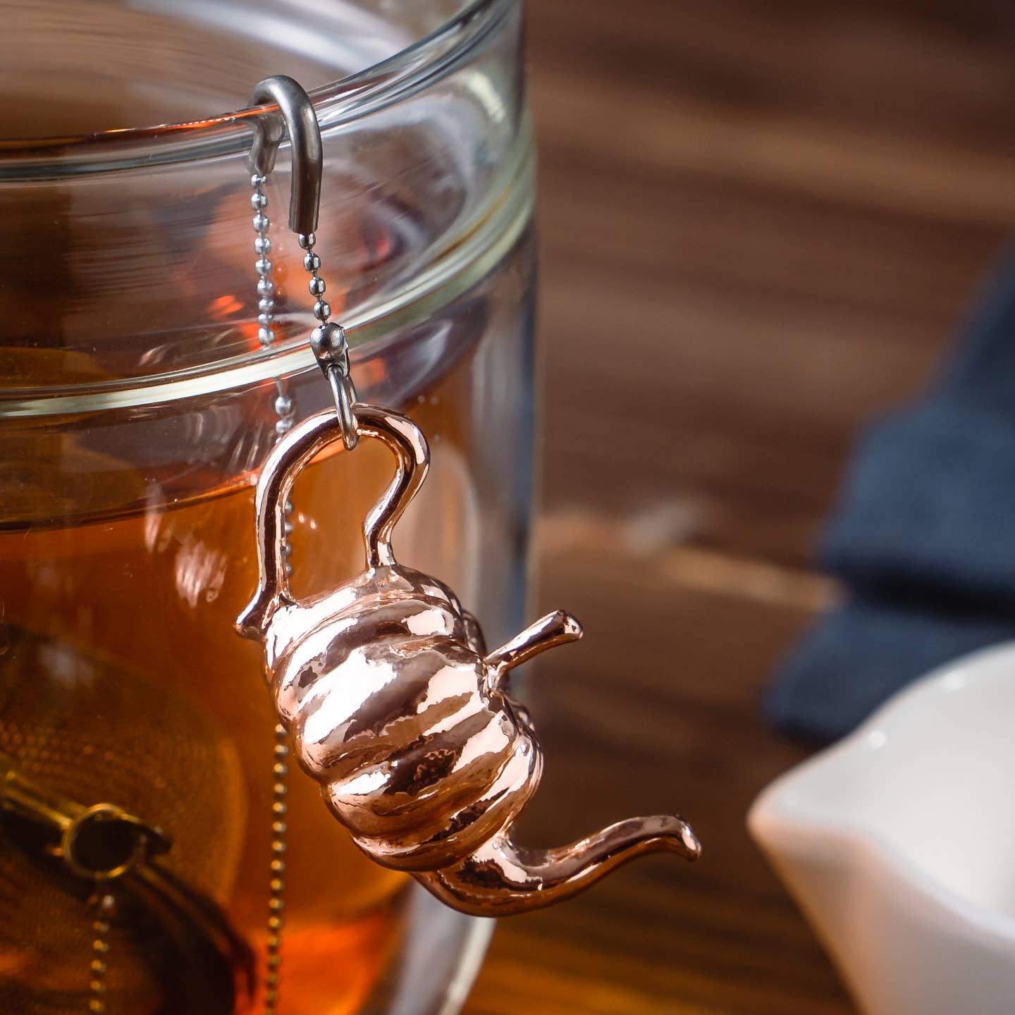 01Living Rose Gold coloured pumpkin-shaped tea infuser ChiChi hangs from the rim of a clear glass mug filled with brewed tea on a wooden table.