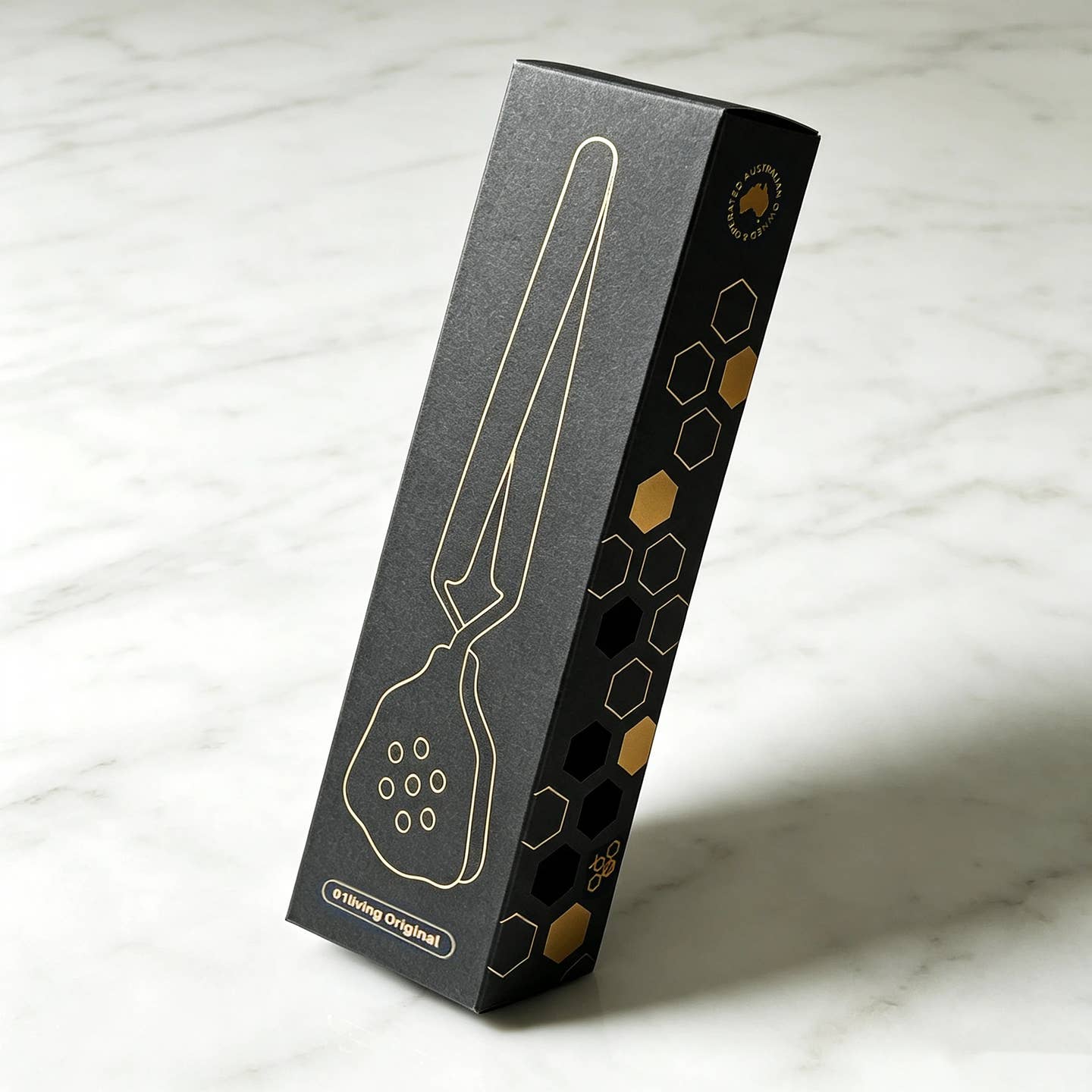 A tall, narrow black product box with gold honeycomb details, standing on a light marble surface of a TenderPress™ Gentle Tea Bag Squeezer.