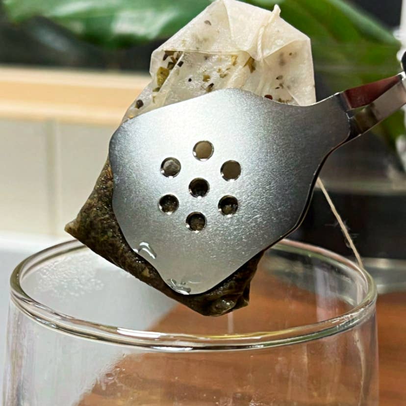 01Living TenderPress™ silver stainless steel tea bag squeezer gently squeezing a tea bag over a teacup to remove excess liquid.