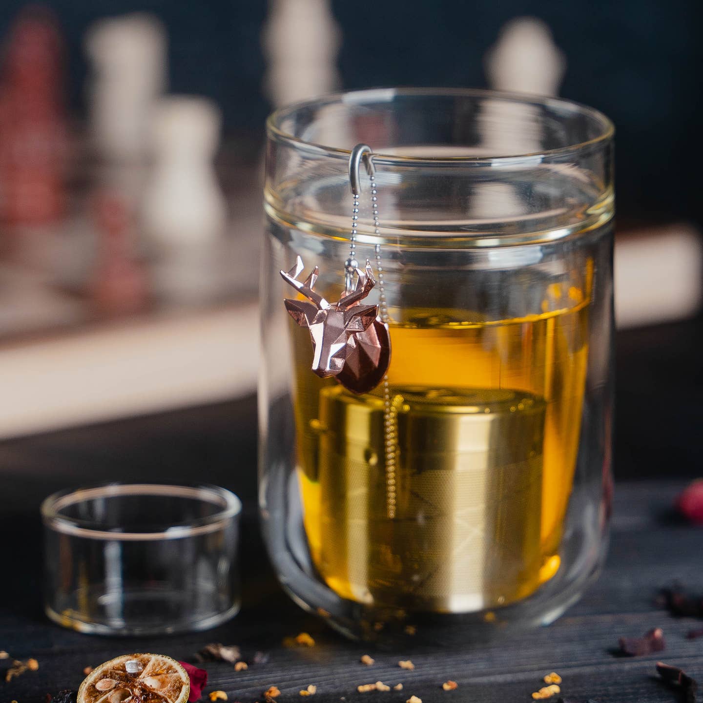 01Living stainless steel tea infuser with a rose gold deer-shaped ChiChi ornament, set on a dark wooden surface with dried tea ingredients and a glass of brewed tea in background