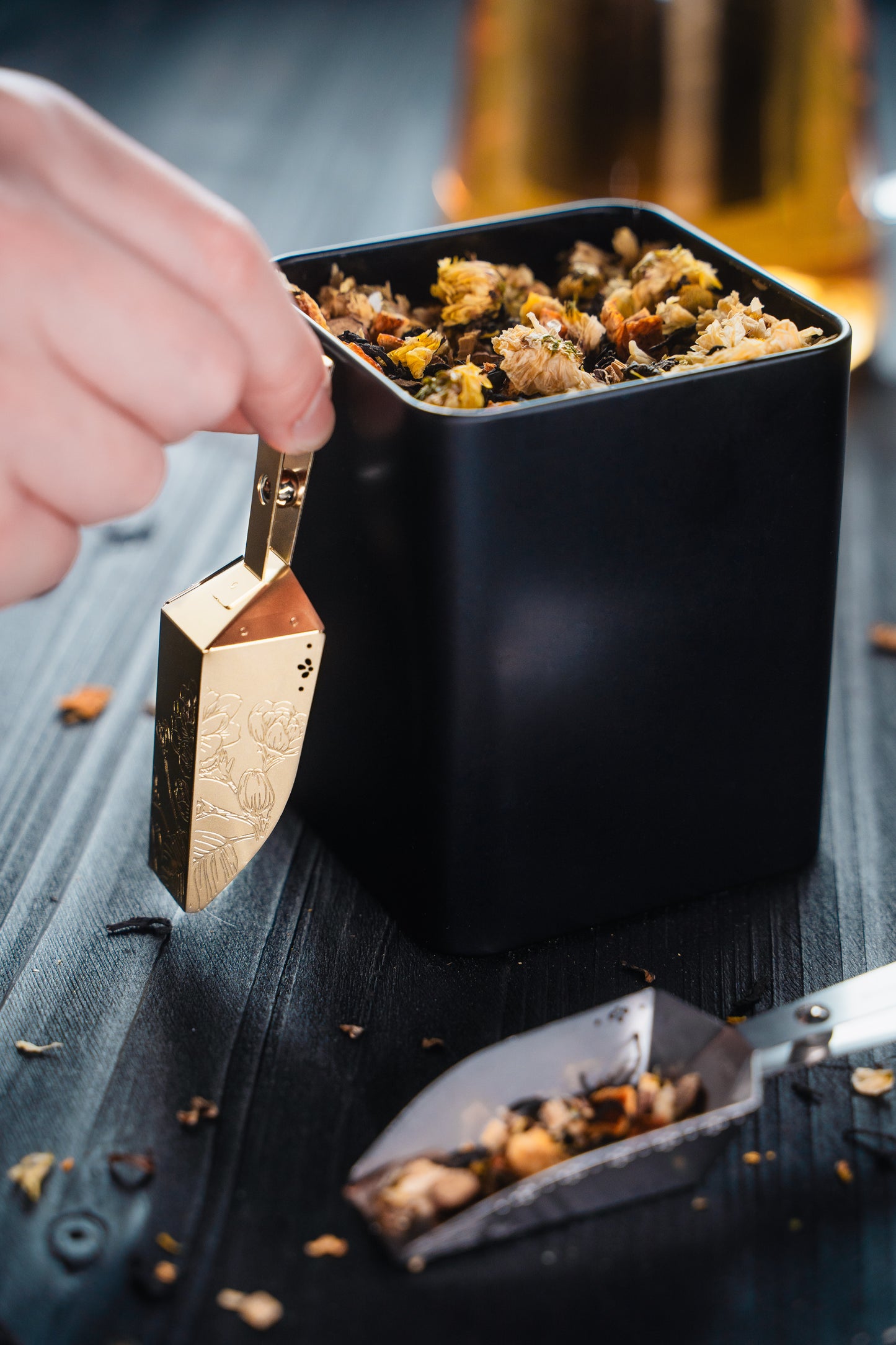 Gold and Silver Magnetic Magic-Scoop™ tea scoops displayed with an open tea tin and loose-leaf tea on a sophisticated black tabletop, with a hand showing how the scoop magnetises to the tin.