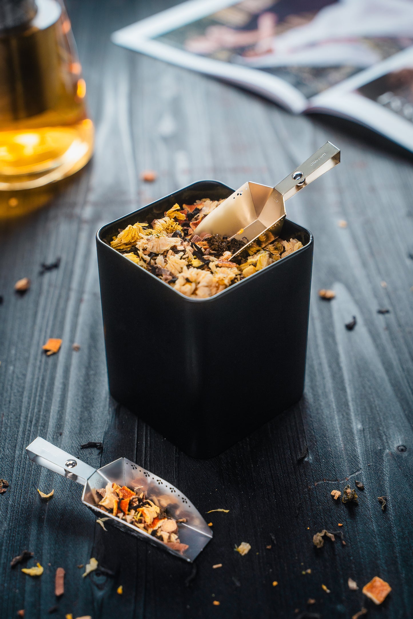 Gold Magnetic Magic-Scoop™ resting within a tea tin of tea with a Silver scoop resting beside it, styled with loose-leaf tea on a black table.