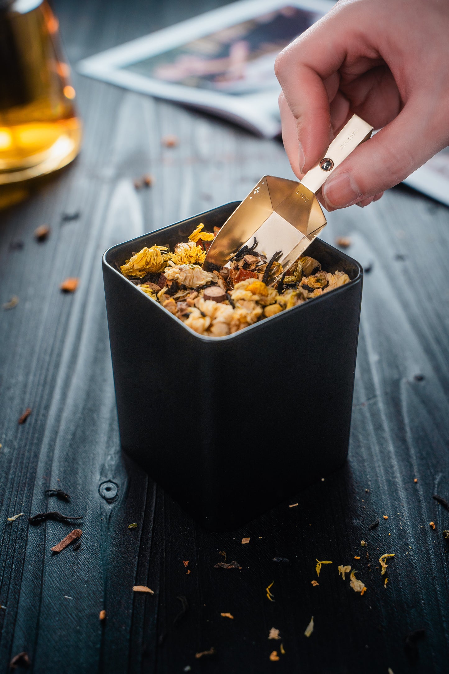 Hand holding the Magnetic Magic-Scoop™ in Gold scooping tea from a tin filled with loose-leaf tea on a black table.