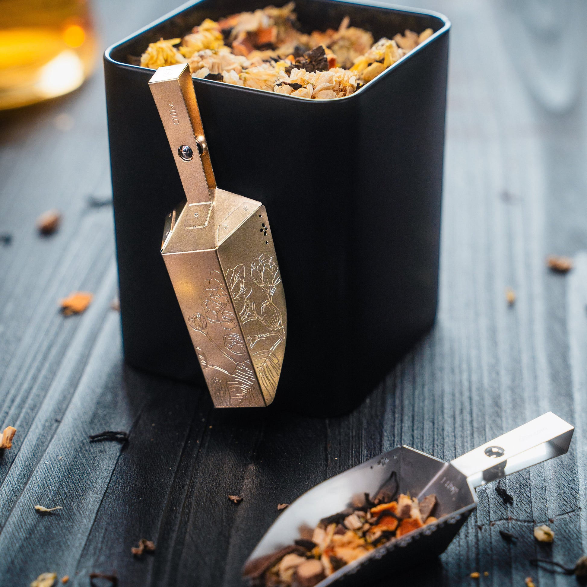 Magnetic Magic-Scoop™ tea scoops in Gold and Silver resting beside an open tea tin filled with loose-leaf tea on a black table.